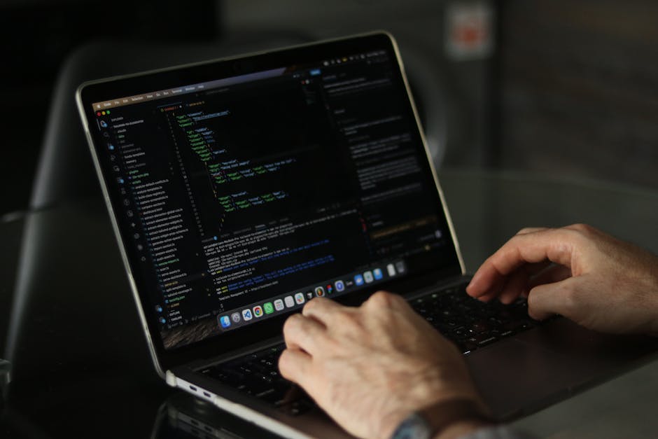 This image shows a person's hands typing on a laptop keyboard, with the screen displaying lines of computer code set against a dark background. The laptop is placed on a smooth, reflective surface, an